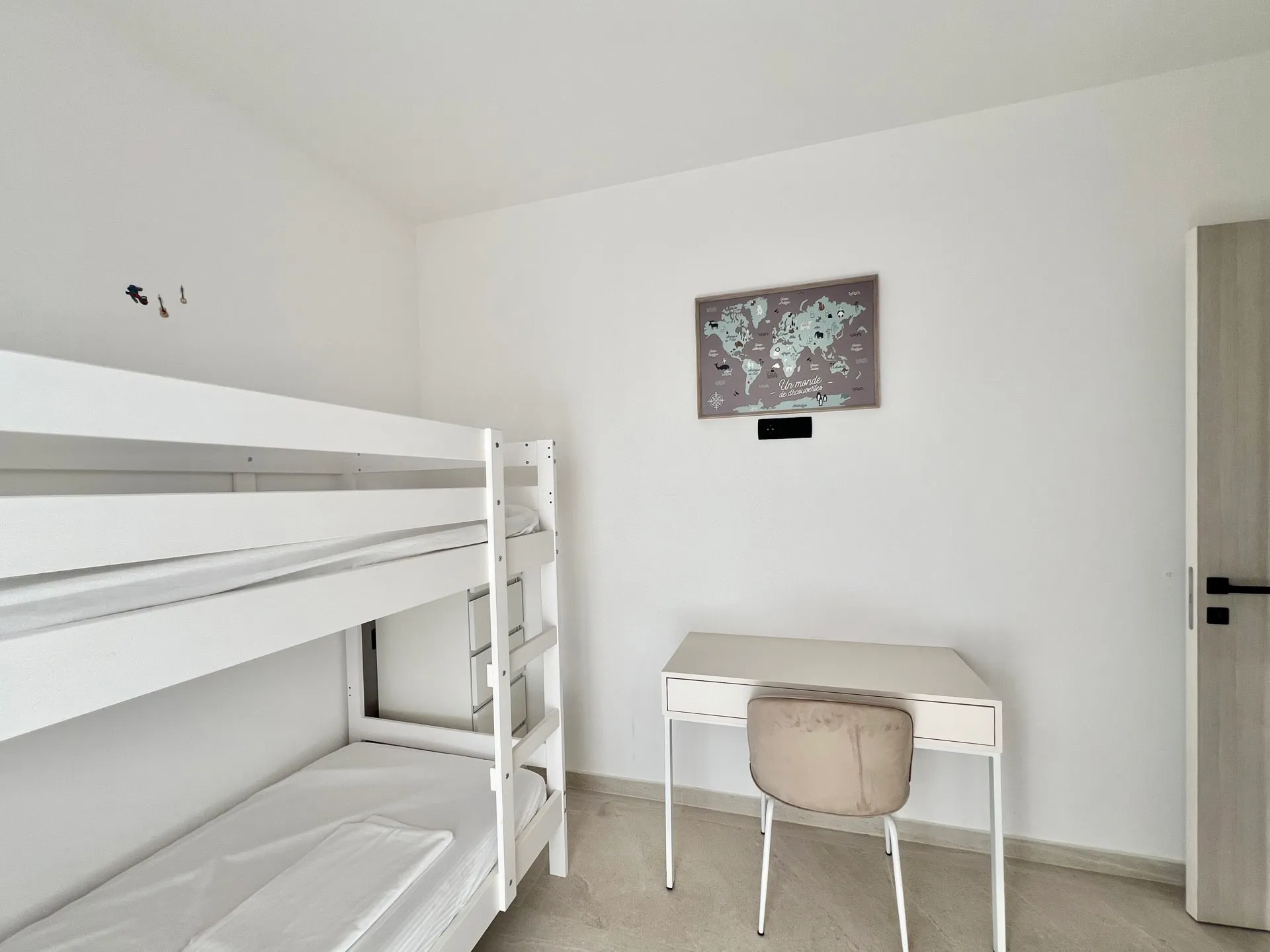 White bunk bed, white desk with a chair, and a framed world map hanging on the plain white wall.
