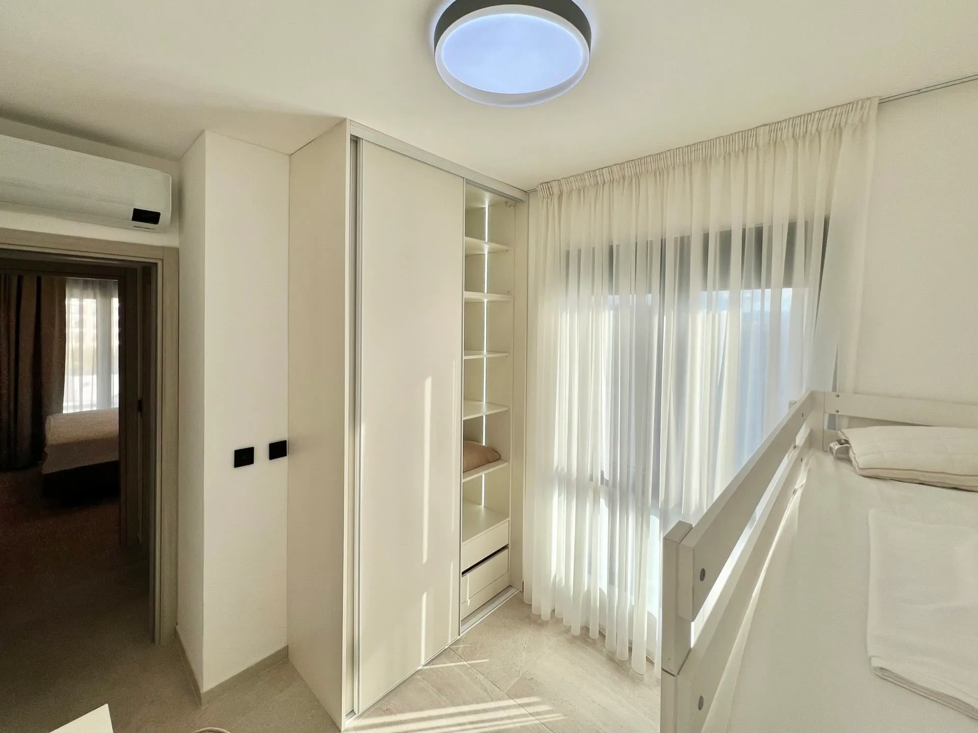 Bright bedroom featuring a white sliding wardrobe, open shelving, and a bed near sheer curtains.
