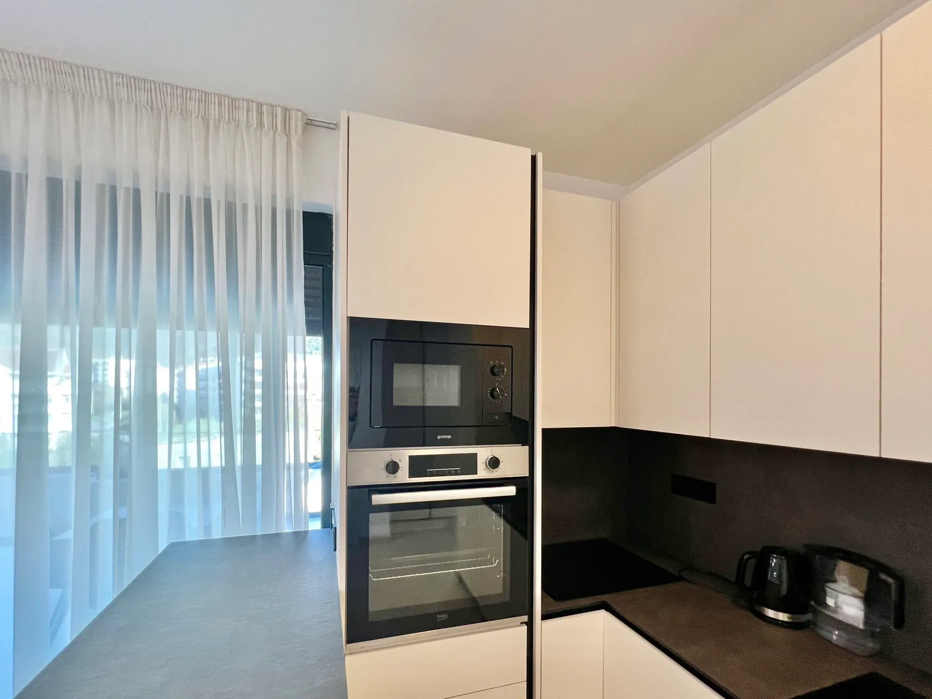 Modern kitchen with white cabinets, built-in oven and microwave, dark countertop, kettle, and sheer curtains near a window.