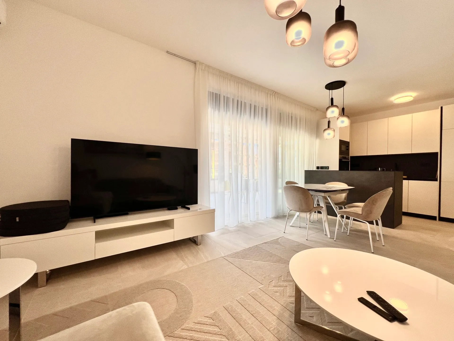 Modern living room featuring a large TV, white coffee table, dining area with chairs, and a kitchen island in the background.