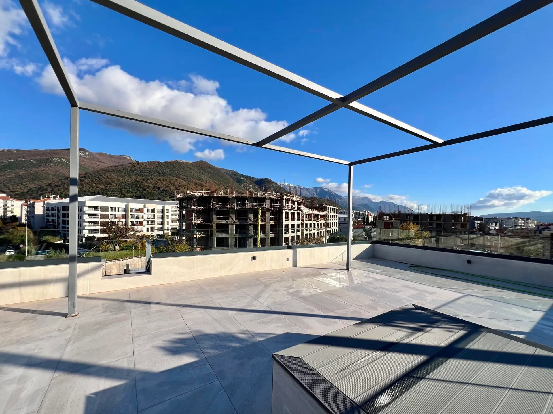 Wide covered balcony with metal frame and tiled floor, overlooking mountains and city buildings.