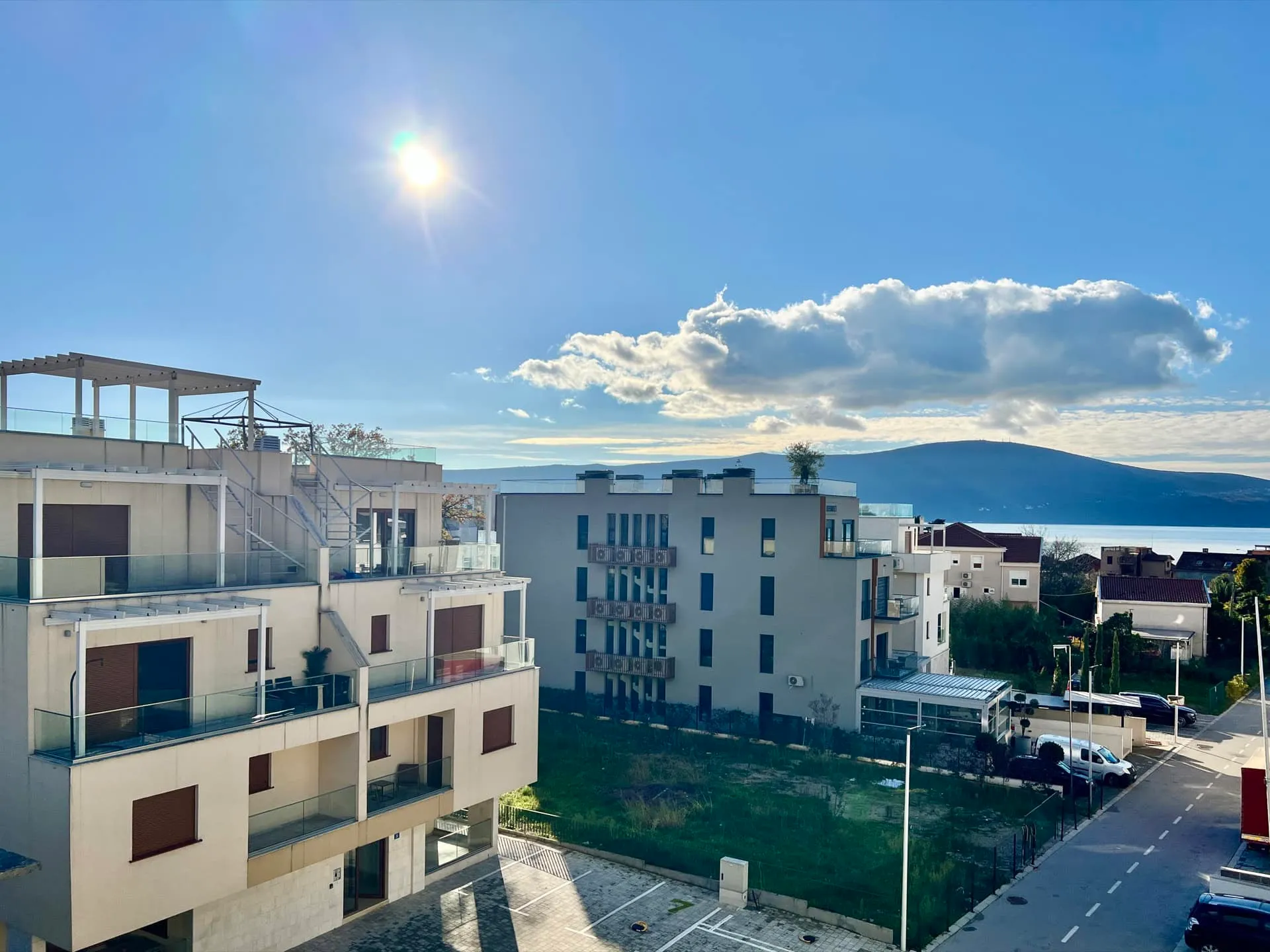 View of modern residential buildings with balconies, a parking area, and a lake with mountains in the background.