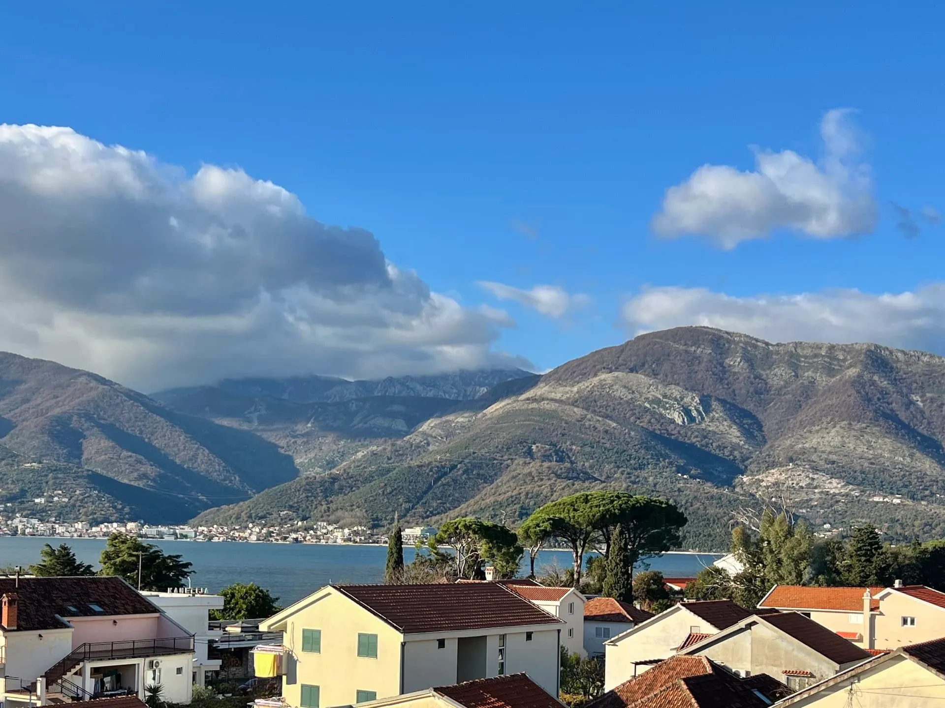 Panoramic view of mountains and water body seen over residential rooftops under a cloudy sky.