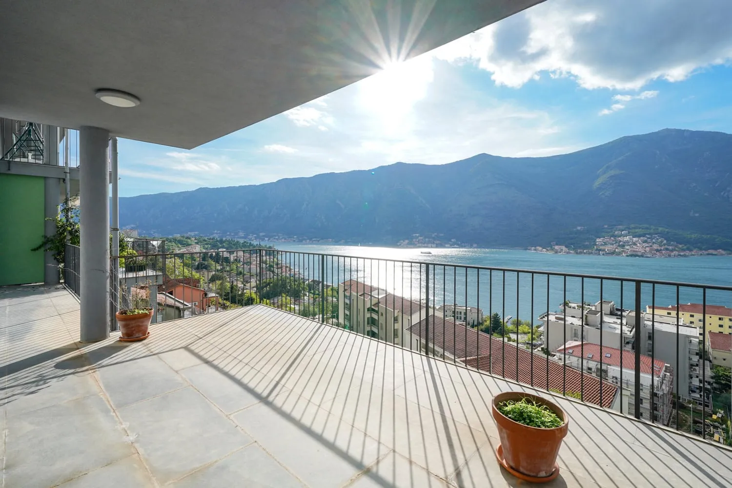 Wide balcony with sea and mountain views, concrete floor, metal railings, and potted plants under bright sun.