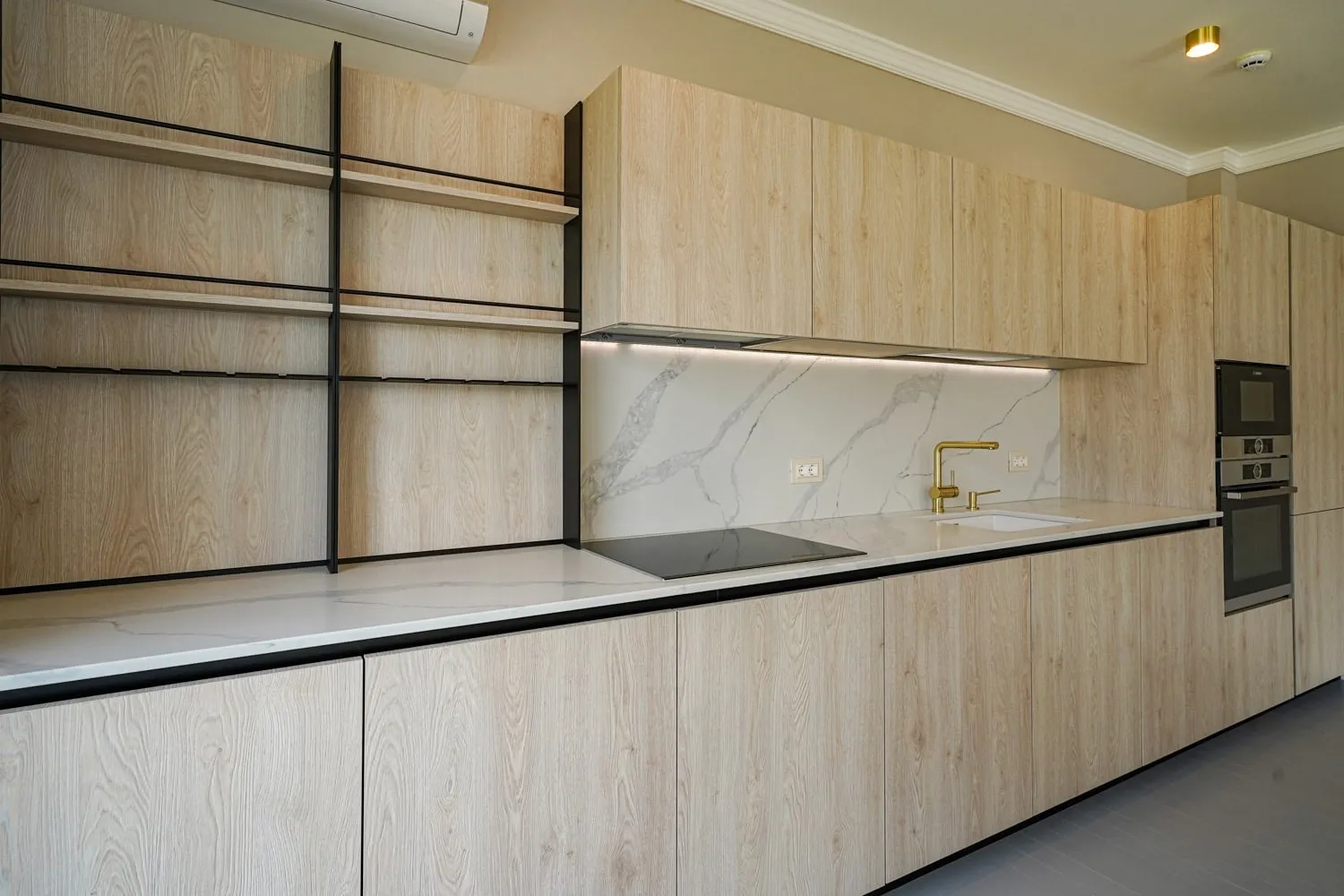 Modern kitchen with light wood cabinets, marble countertop, gold faucet, induction cooktop, and built-in ovens.