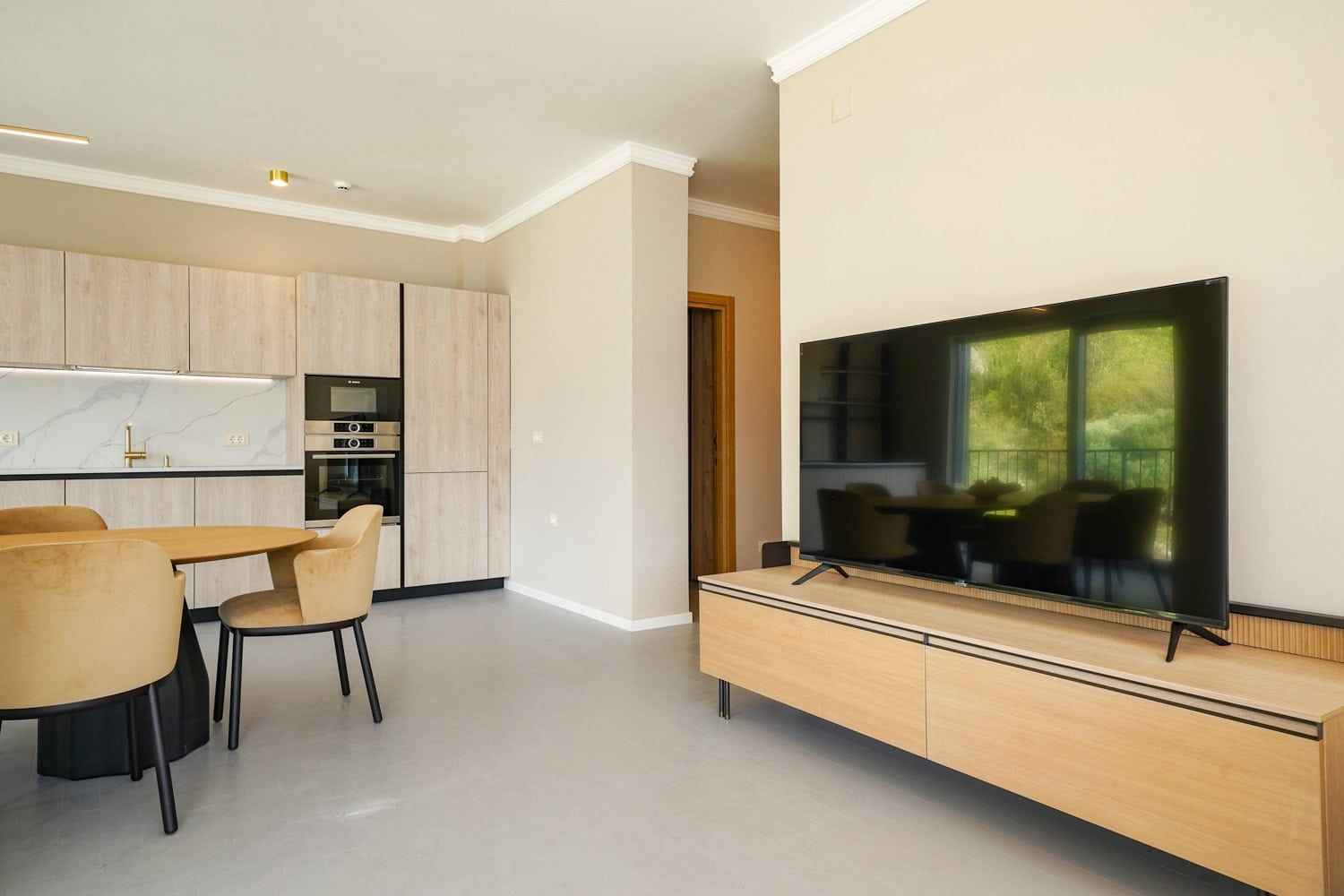 Modern living room with kitchen in an apartment in Dobrota, Montenegro: light furniture, TV area, and a view of greenery from the window.