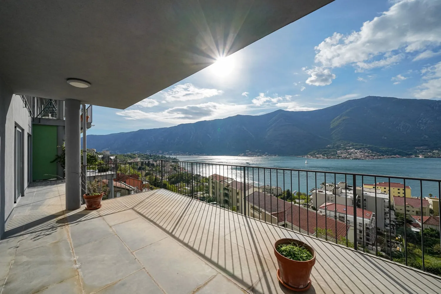 Wide stone balcony with metal railing overlooking the sea and mountains under bright sun.