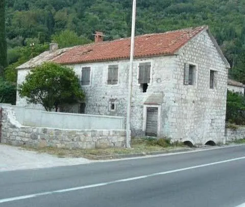 Old stone house with red tiled roof viewed from outside next to a road against a forested hill background.