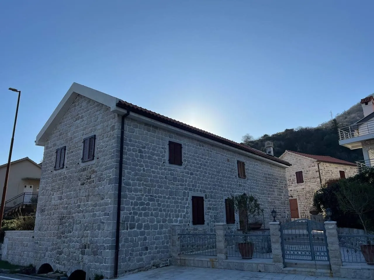 Exterior view of a stone house with wooden shutters, tiled roof, and wrought iron fence against a mountain backdrop.