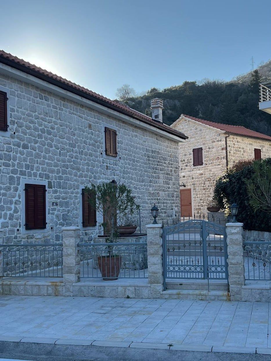 Stone house exterior with wooden shutters, wrought iron gate, and fence against a mountain backdrop.