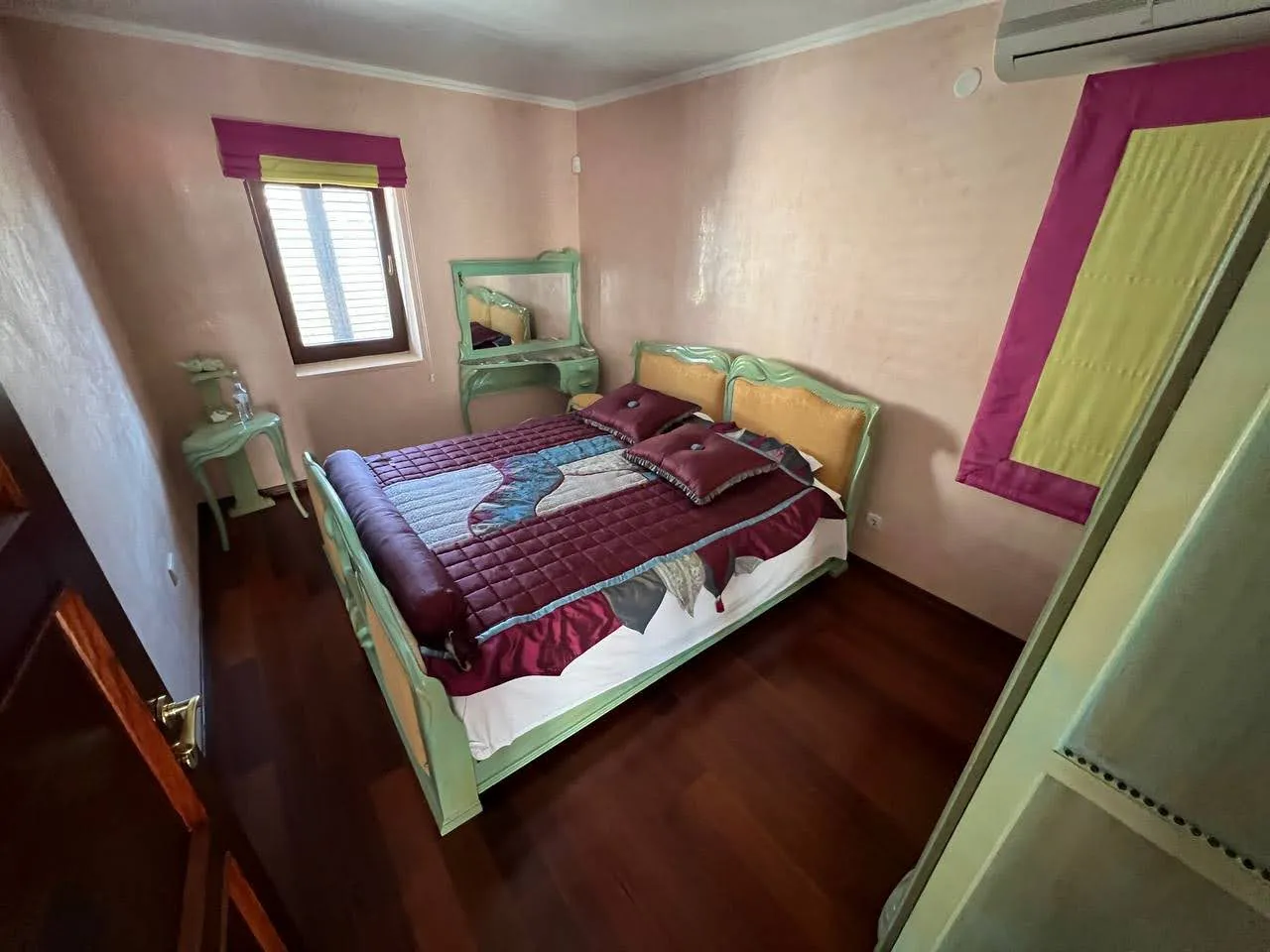 Bedroom featuring a double bed with a green frame, pink walls, dark wood floors, and a vanity table near the window.