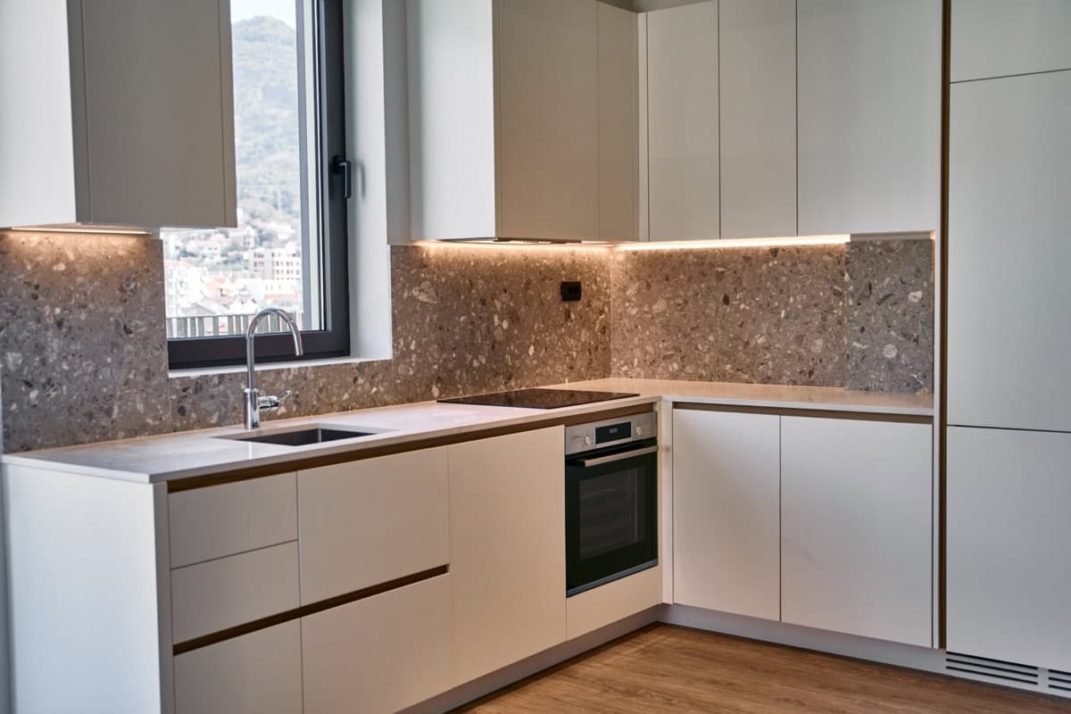 Modern kitchen in an apartment in Tivat, Montenegro: white cabinetry, terrazzo backsplash, and mountain views from the window.