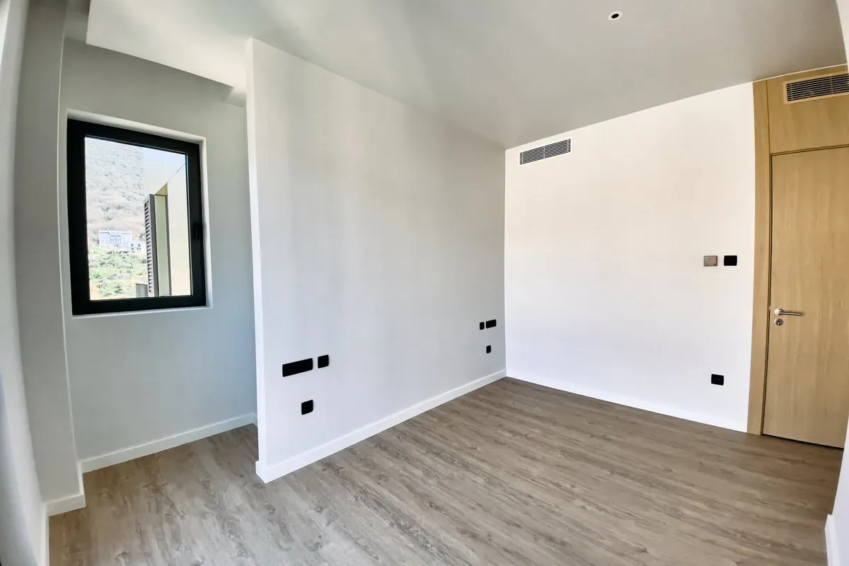 Empty room with white walls, wood flooring, a window, and a door. Visible electrical outlets and vents.