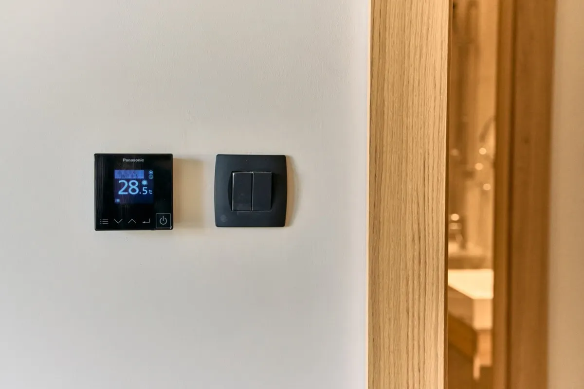Digital Panasonic thermostat and light switch on a white wall next to a wooden door frame.