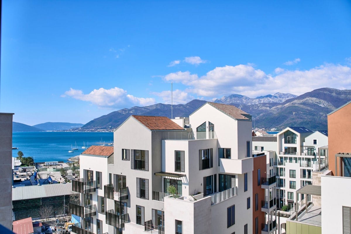 View of modern residential buildings in Tivat, Montenegro, with a panoramic sea view and mountain scenery on a sunny day.