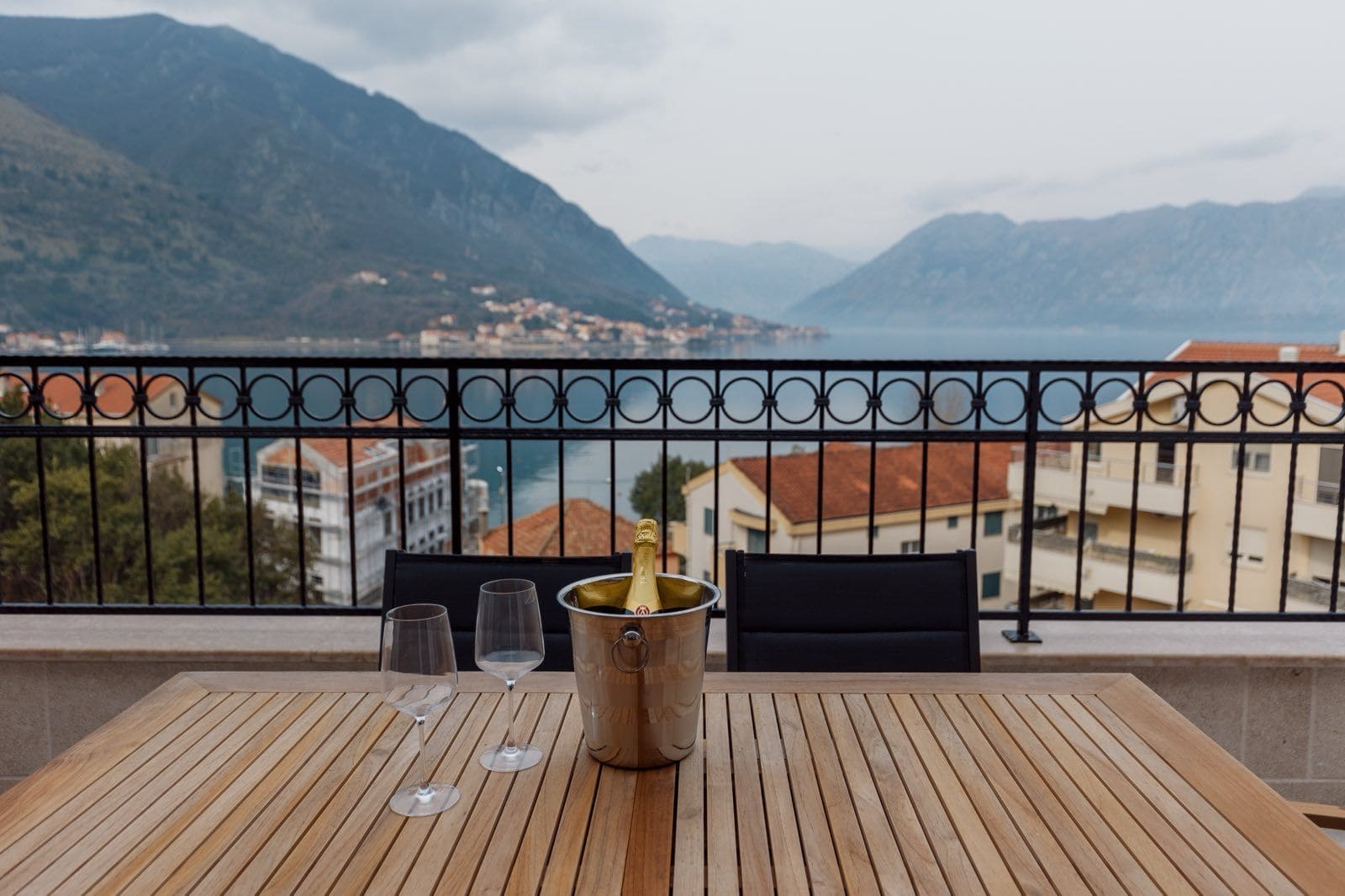 Wooden table with glasses on the apartment balcony in Dobrota, Montenegro, with panoramic views of the Bay of Kotor and the mountains.