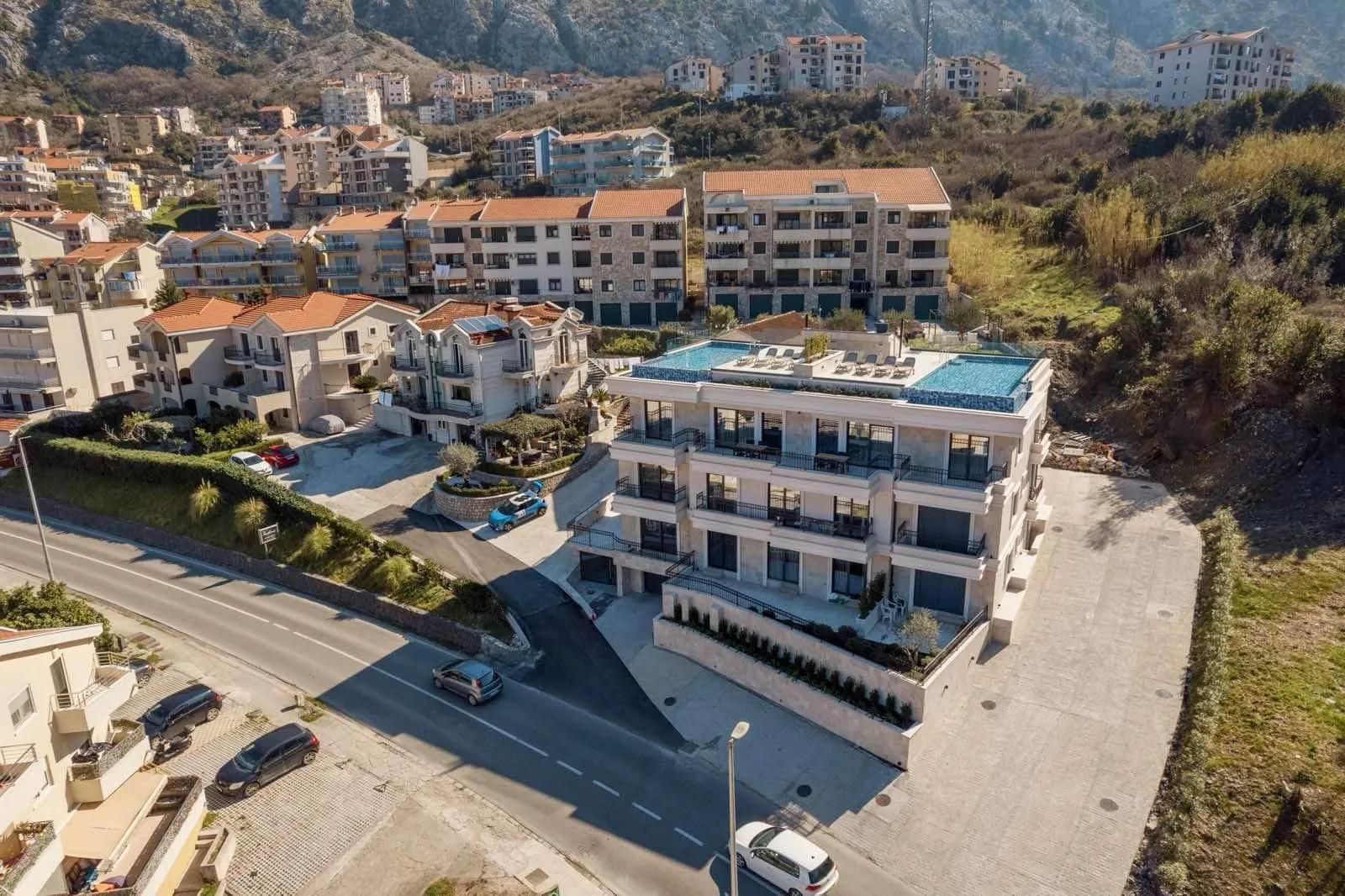 Aerial view of a residential complex featuring a rooftop pool, adjacent road, and mountain backdrop.