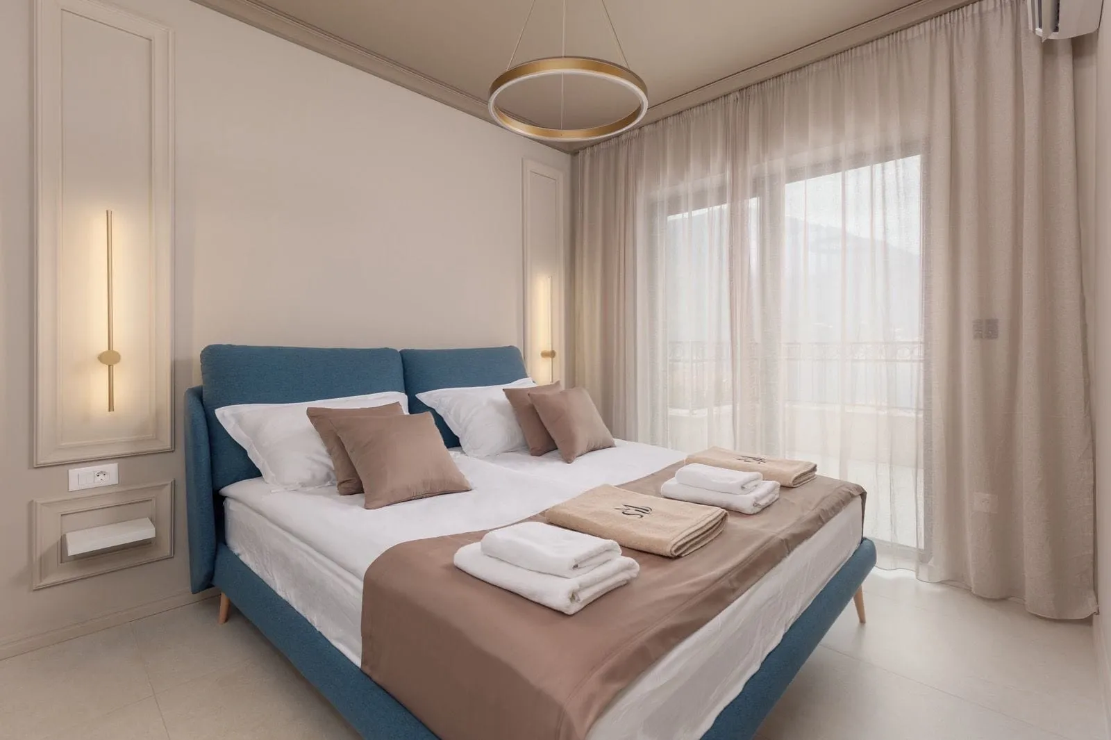Modern bedroom featuring a blue bed frame, white linens, folded towels, sheer curtains, and a circular ceiling light.