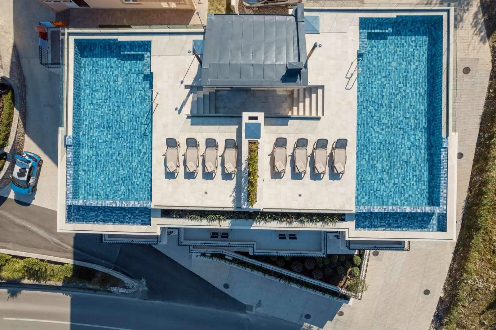 Aerial view of two blue tiled swimming pools flanking a central patio with lounge chairs.