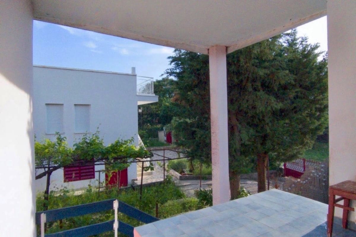 View from the covered terrace of the garden and neighboring house in Dobra Voda, Montenegro: tiled floor and a green plot with trees.