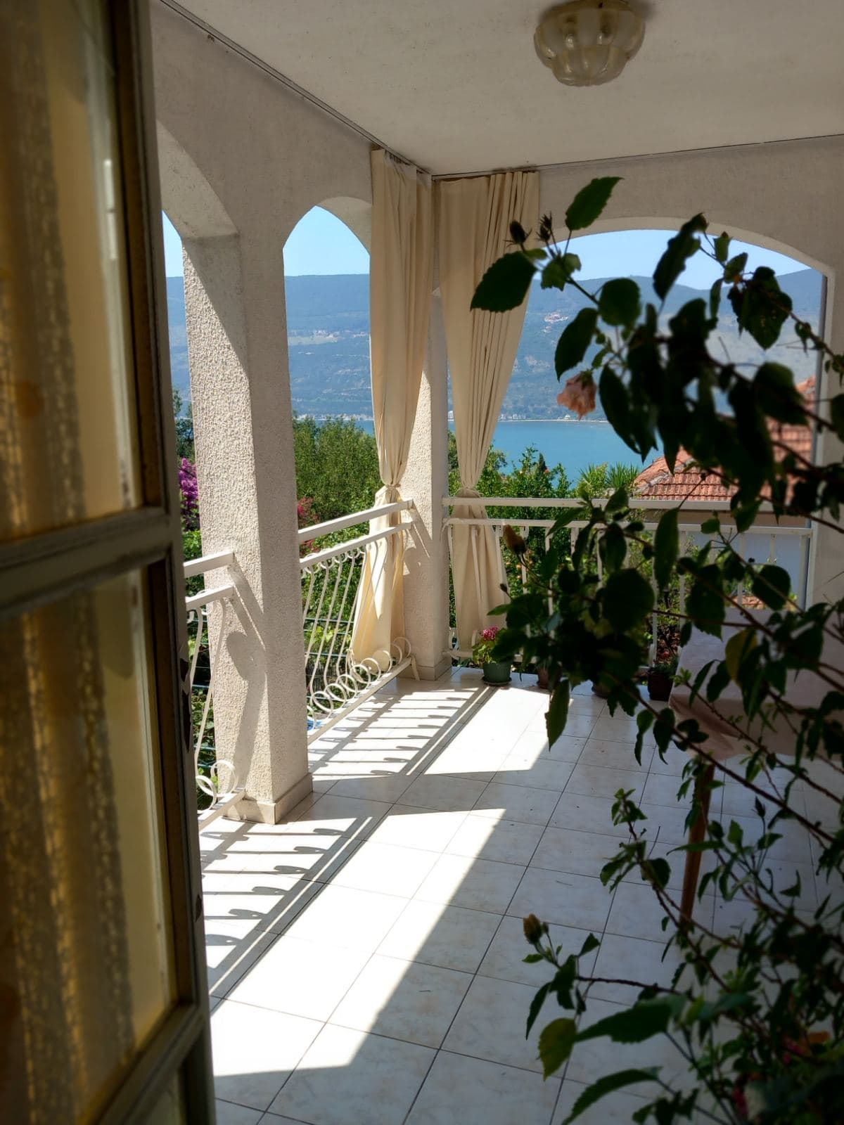 Spacious balcony with arched openings and sea view in a house in Igalo, Montenegro, with tiled floor and light curtains.