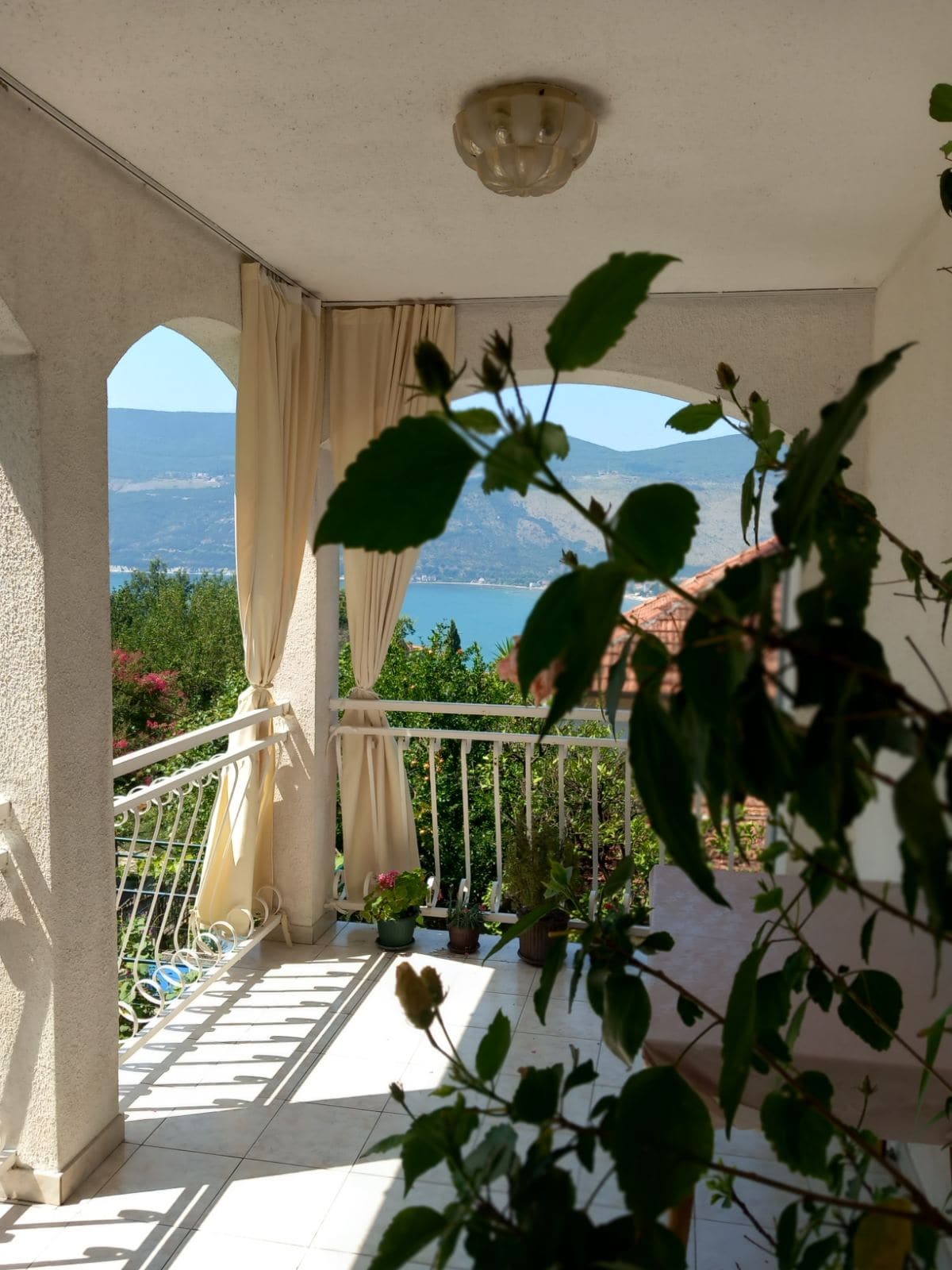 Balcony with sea and mountain views in a house in Igalo, Montenegro: arched openings, light curtains, and houseplants.