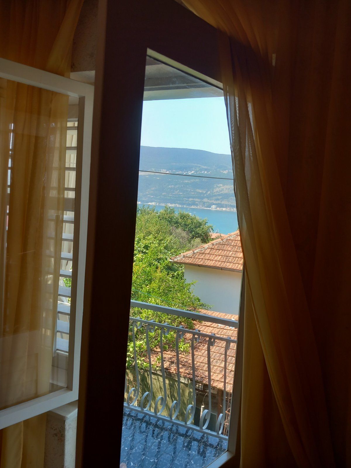View from the bedroom window of the sea and mountains in a house in Igalo, Montenegro: a balcony with wrought-iron railing and yellow curtains.