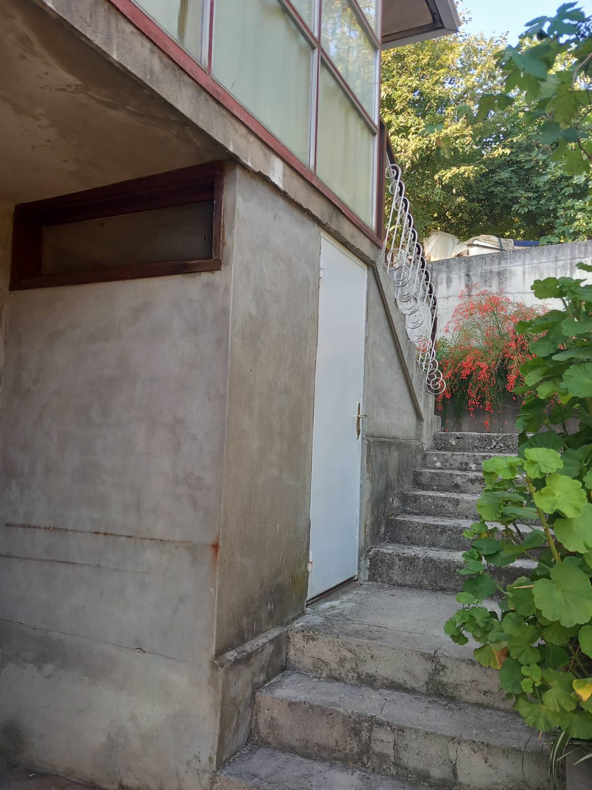 Entrance to a house in Igalo, Montenegro: concrete stairs, a white door, wrought-iron railings, and flowering plants in the yard.