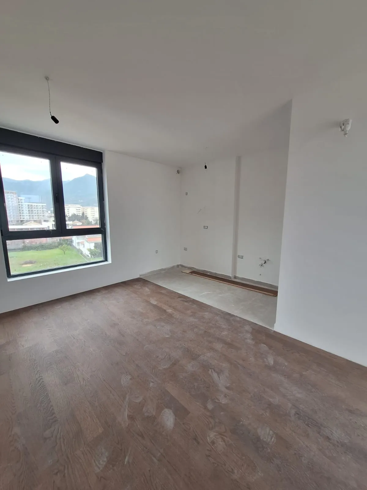Empty room with wood flooring and large window overlooking city and mountains, under renovation.