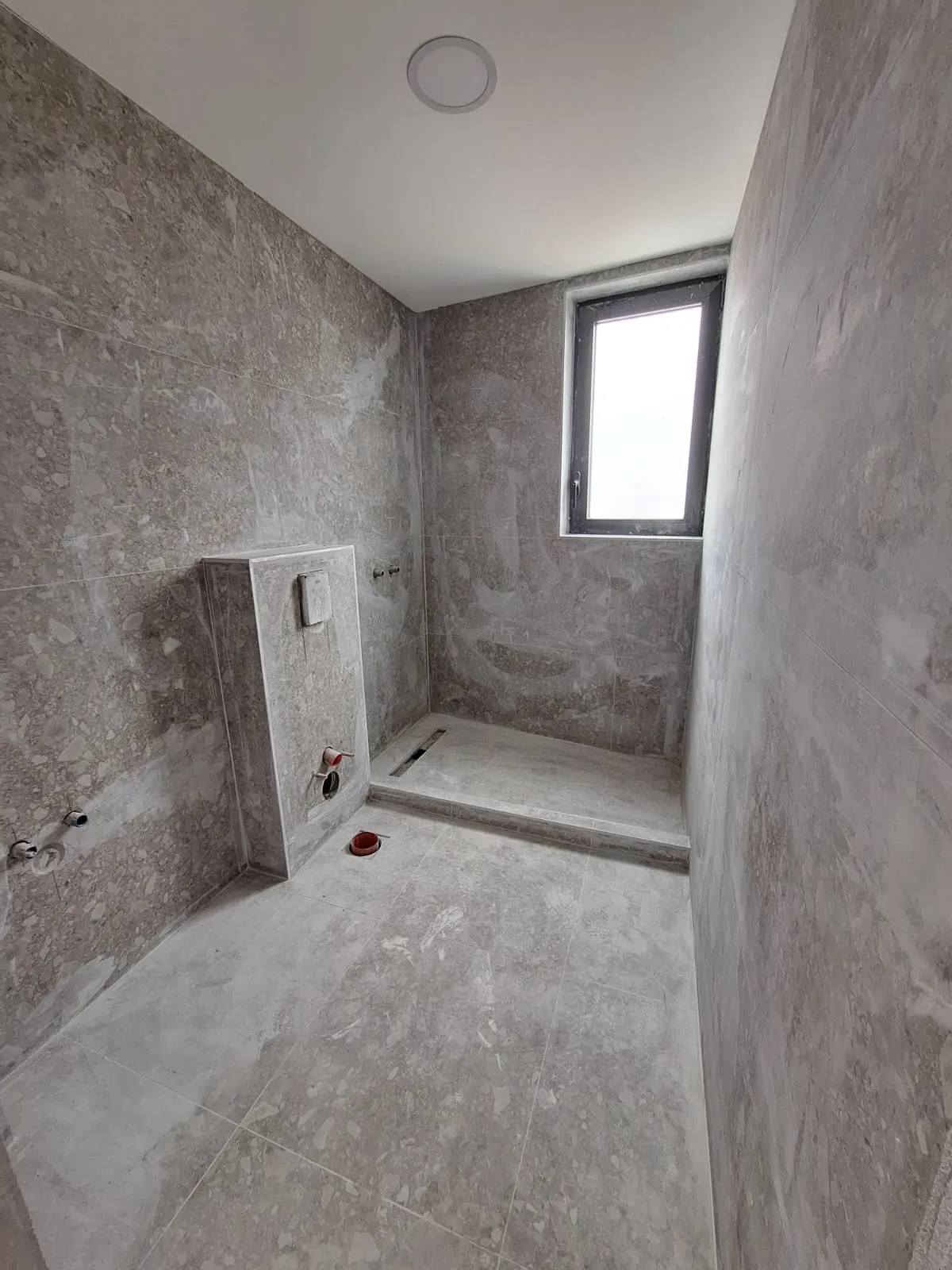 Unfinished bathroom with gray terrazzo tiles, a raised shower base, and exposed plumbing pipes.