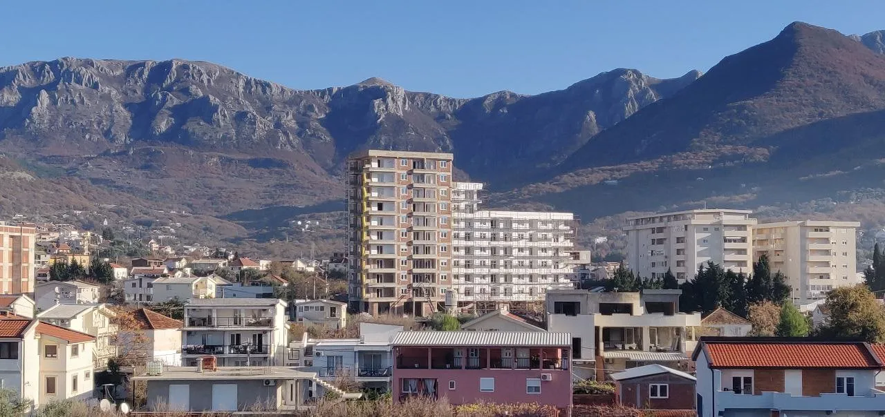 Urban landscape featuring multi-story buildings and houses set against a backdrop of mountains under a clear sky.