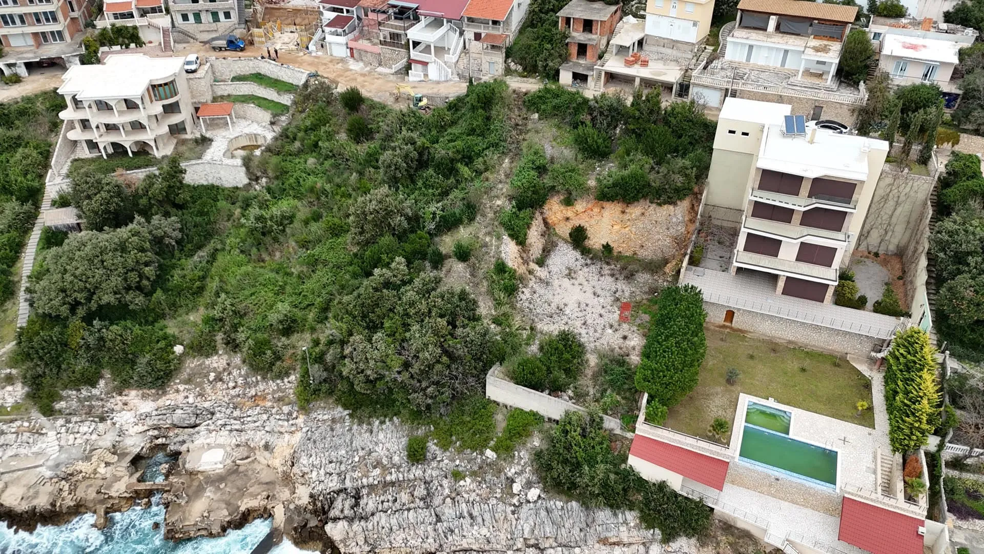 Aerial view of a coastal plot with houses, dense vegetation, and a rocky shoreline meeting the sea.