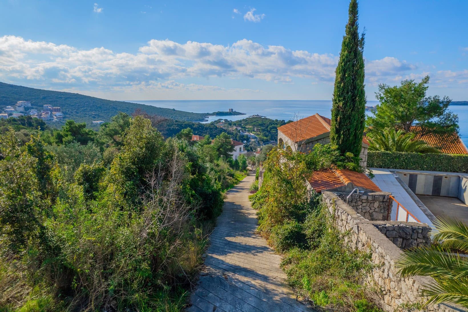 Land plot in Žanjice, Montenegro: panoramic view of the sea, hills, and Mediterranean nature with a path leading to the house.