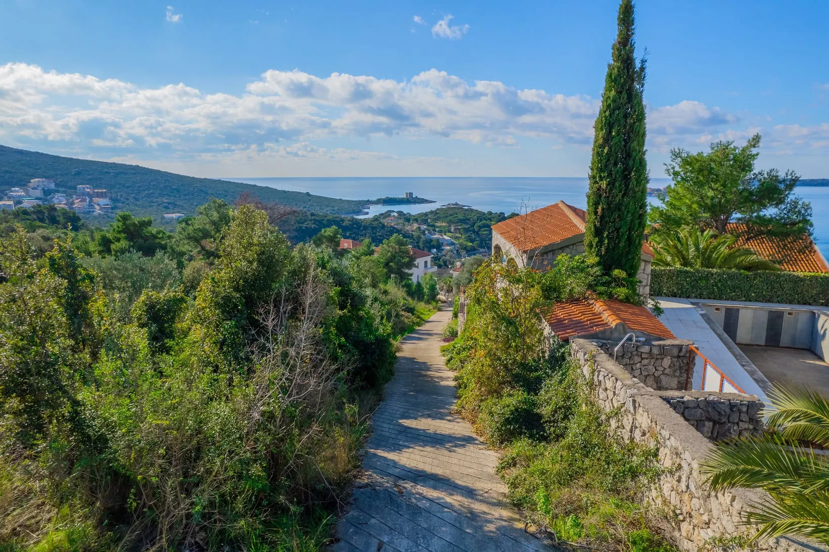 Scenic coastal view featuring a stone path, walls, and tiled roofs amidst lush greenery.