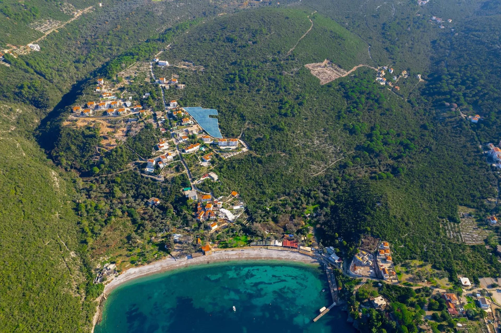 Aerial view of a coastal resort featuring a large pool, beach, and buildings on a green hillside.