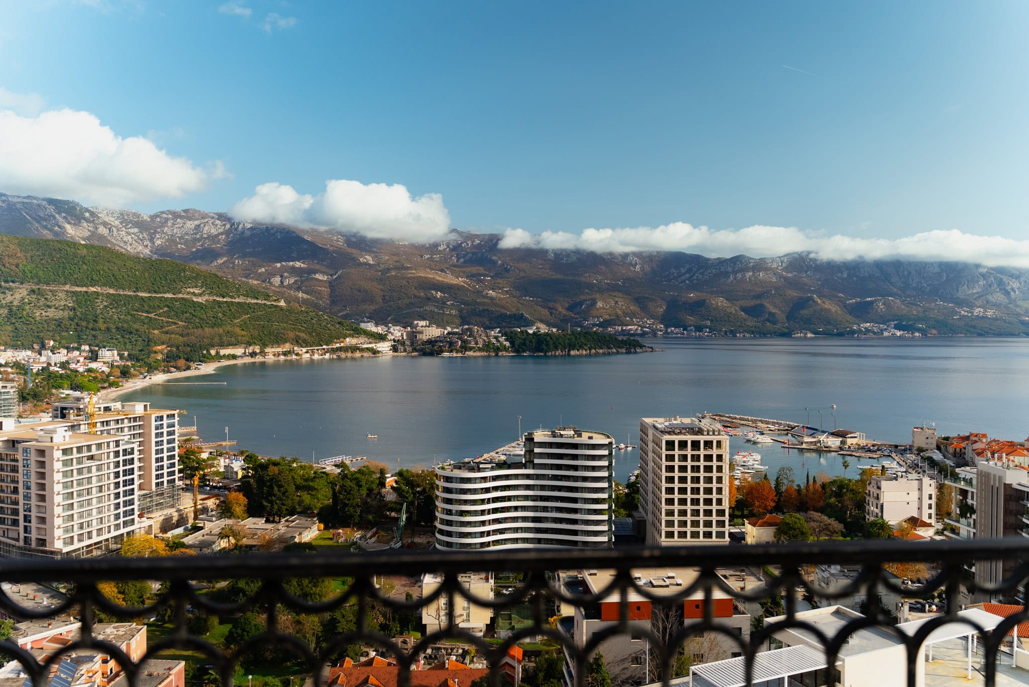 Panoramic view from the balcony of the sea, mountains, and the city of Budva, Montenegro, with a wrought-iron railing in the foreground.