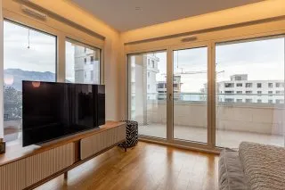 Living room with TV on a wooden console, hardwood floors, and large windows overlooking a balcony.