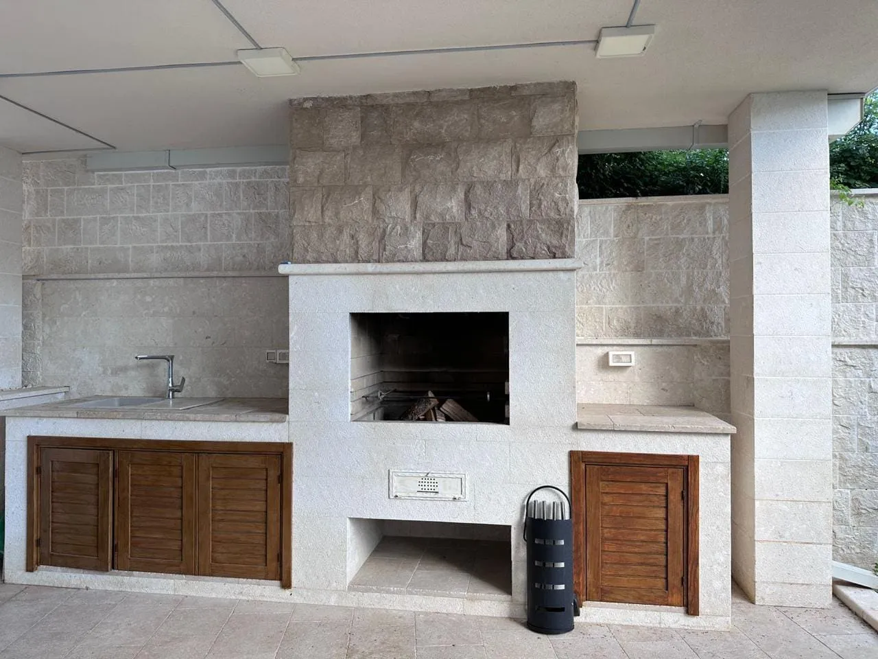 Outdoor kitchen featuring a stone barbecue grill, sink, and wooden cabinets under a covered patio.