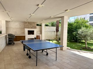Covered terrace with a barbecue area and a ping pong table at a villa in Bečići, Montenegro, with access to a green garden.