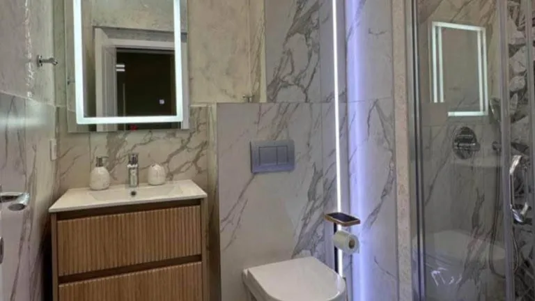 Bathroom with marble tiles, wooden vanity sink, toilet, and glass shower enclosure.