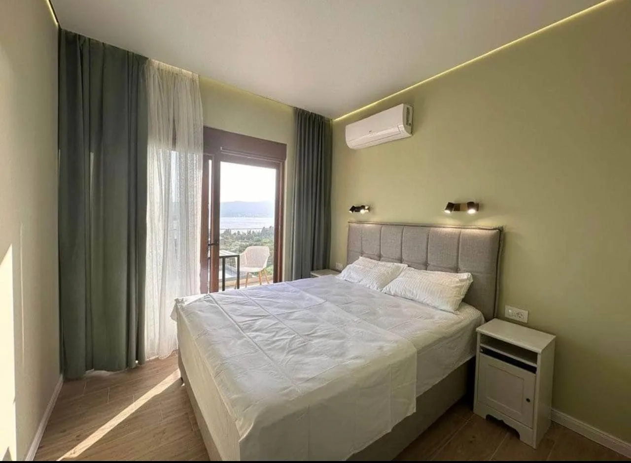 Bright bedroom featuring a double bed with white linens, wall-mounted AC unit, and a balcony offering a scenic water view.