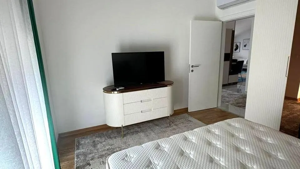 A mattress is visible in the foreground, a TV sits on a white cabinet with a wood top, next to an open door.