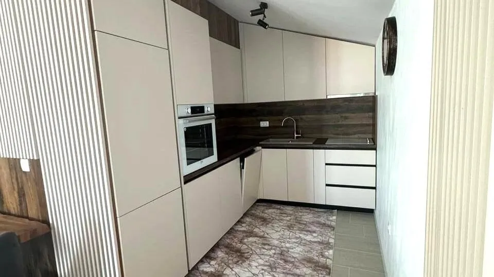 Modern kitchen with beige cabinets, built-in oven, sink, dark countertop, and wood-effect backsplash wall.