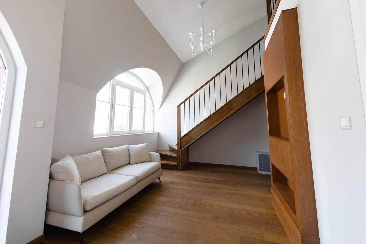 Living room in an apartment in Tivat, Montenegro: light-colored sofa, wooden staircase, arched window, and parquet floor.