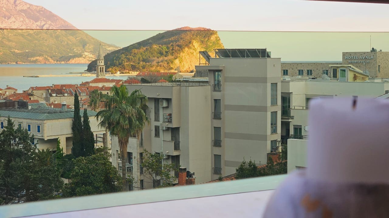 View from the apartment balcony in Budva, Montenegro: a panorama of the Old Town, the Avala Hotel, the mountains, and the Adriatic Sea.