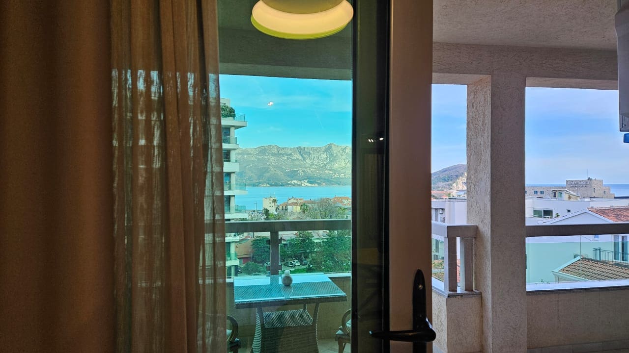 View from the apartment balcony in Budva, Montenegro: wicker furniture, panoramic sea and mountain views, modern finishes.