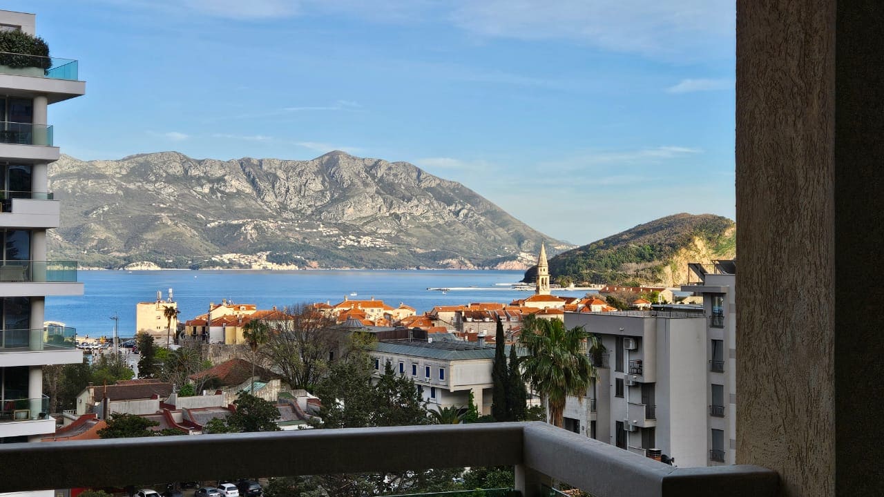 View from the apartment balcony in Budva, Montenegro: a panorama of the Adriatic Sea, mountains, and the Old Town with terracotta roofs.