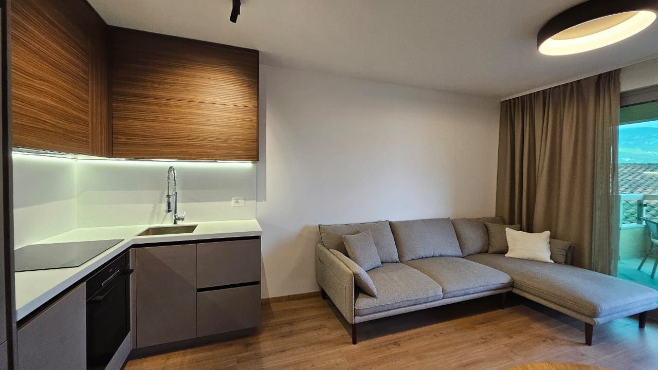 Modern living room with kitchen in an apartment in Budva, Montenegro: gray corner sofa, built-in furniture, and a view of the terrace.