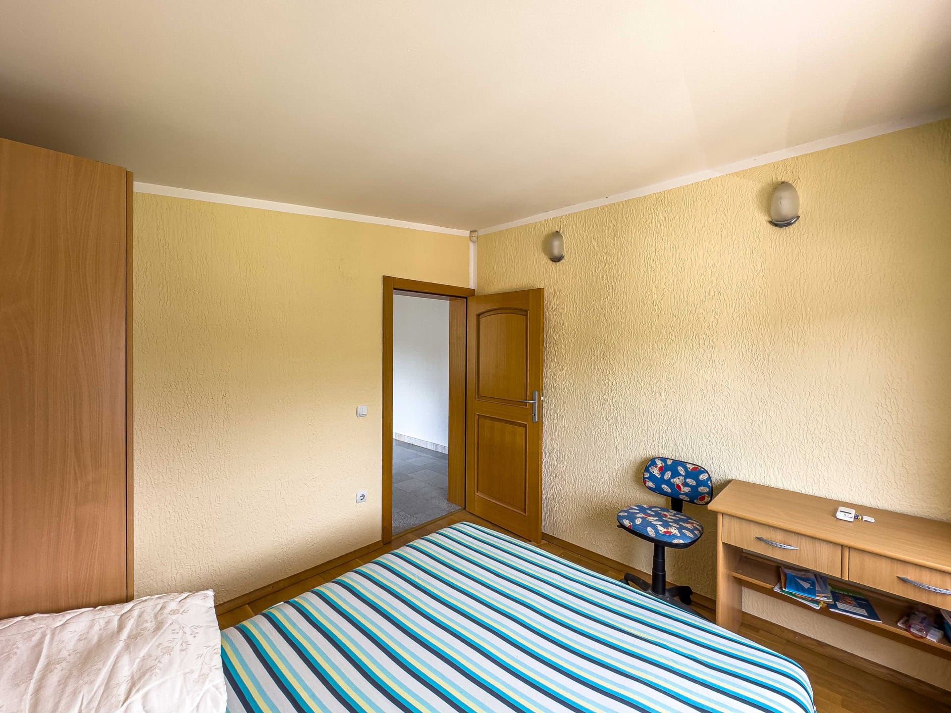 Bedroom in a house in Utjeha, Montenegro: bed with a striped bedspread, wooden table, chair and wardrobe, yellow walls.