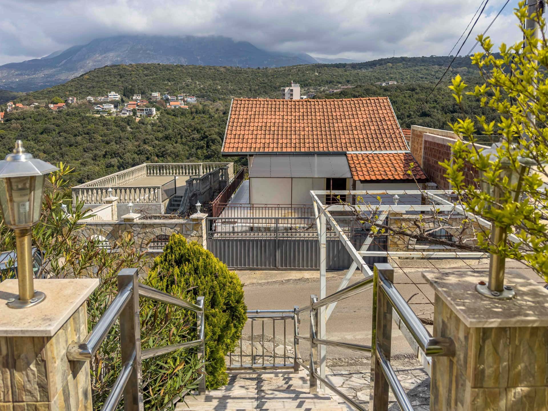 View of a house with a tiled roof and terrace in the village of Uteha, Montenegro, against a backdrop of mountains and green landscape.